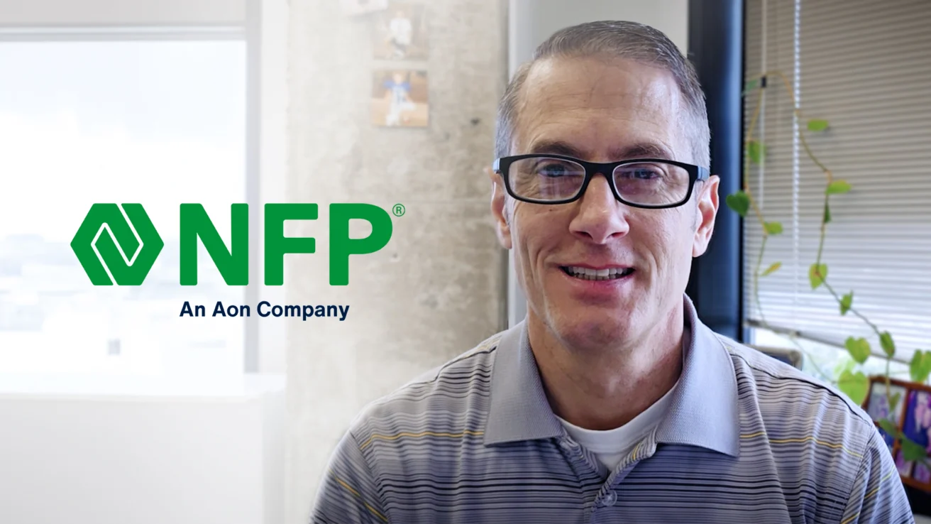 A man wearing glasses and a striped polo shirt sits in an office with the NFP, An Aon Company, logo visible on the wall beside him.