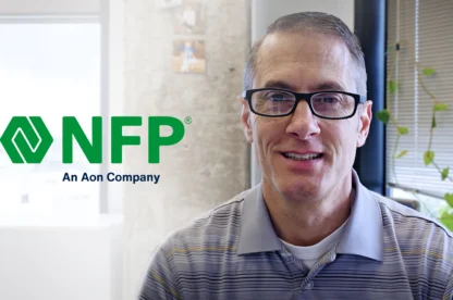 A man wearing glasses and a striped polo shirt sits in an office with the NFP, An Aon Company, logo visible on the wall beside him.