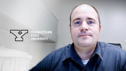 A man in a collared shirt sits in an office. The Youngstown State University logo and name are displayed on the wall beside him.