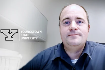 A man in a collared shirt sits in an office. The Youngstown State University logo and name are displayed on the wall beside him.