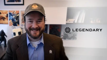 A man in a brown blazer and cap smiles in an office with Legendary Entertainment logo on the wall and movie posters behind him.