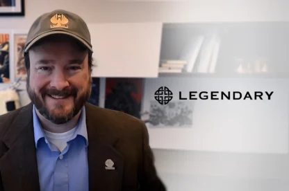 A man in a brown blazer and cap smiles in an office with Legendary Entertainment logo on the wall and movie posters behind him.