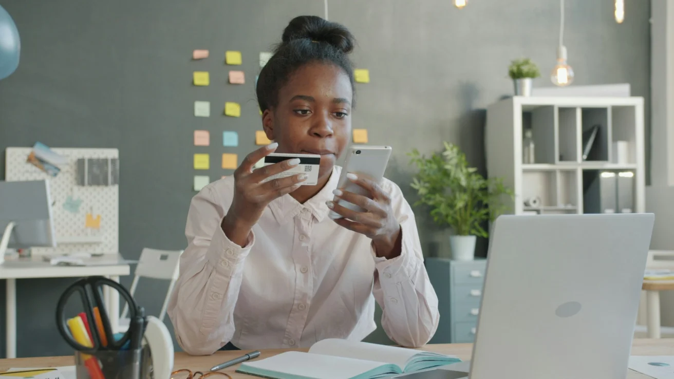 Employee holding a credit card and smartphone while working on a laptop, representing safe online behavior.
