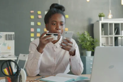 Employee holding a credit card and smartphone while working on a laptop, representing safe online behavior.