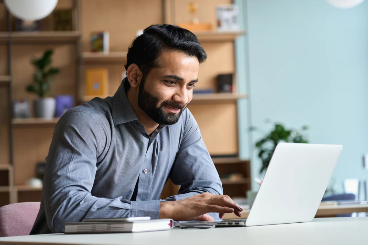 Man in a gray shirt sits at a desk, smiling while using a laptop, with notebooks and shelves in the background.