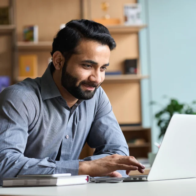 Man in a gray shirt sits at a desk, smiling while using a laptop, with notebooks and shelves in the background.