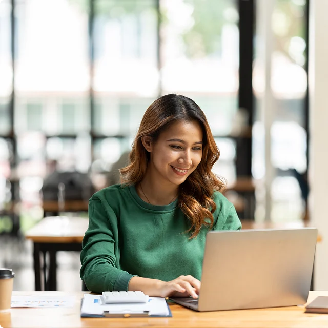 A woman in a green sweater is smiling while working on a laptop at a table with papers and a coffee cup in a bright, modern workspace.