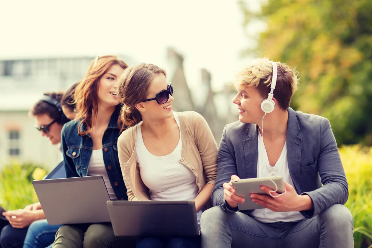 Four young adults sit outdoors with laptops and a tablet, wearing casual clothing and headphones, engaged in conversation and using their devices. Trees and greenery are in the background.
