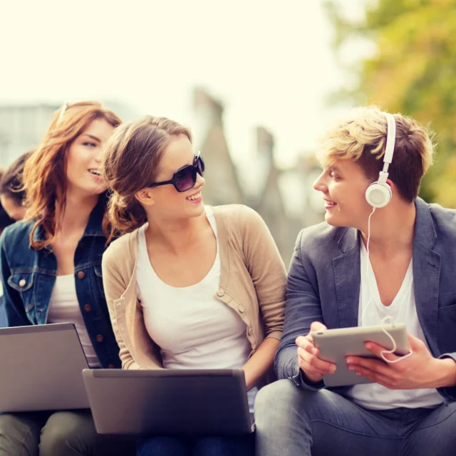 Four young adults sit outdoors with laptops and a tablet, wearing casual clothing and headphones, engaged in conversation and using their devices. Trees and greenery are in the background.