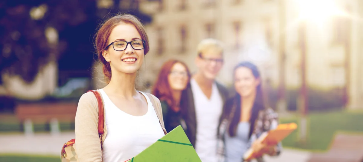 A young woman with glasses holds folders and smiles at the camera, with three other young adults standing and talking in the blurred background outdoors.