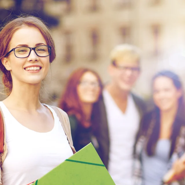A young woman with glasses holds folders and smiles at the camera, with three other young adults standing and talking in the blurred background outdoors.