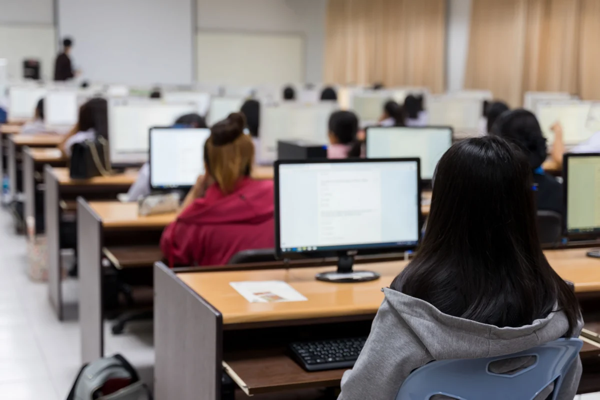 Students sit at desks with computers in a classroom, facing a whiteboard and an instructor at the front of the room.