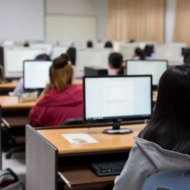 Students sit at desks with computers in a classroom, facing a whiteboard and an instructor at the front of the room.