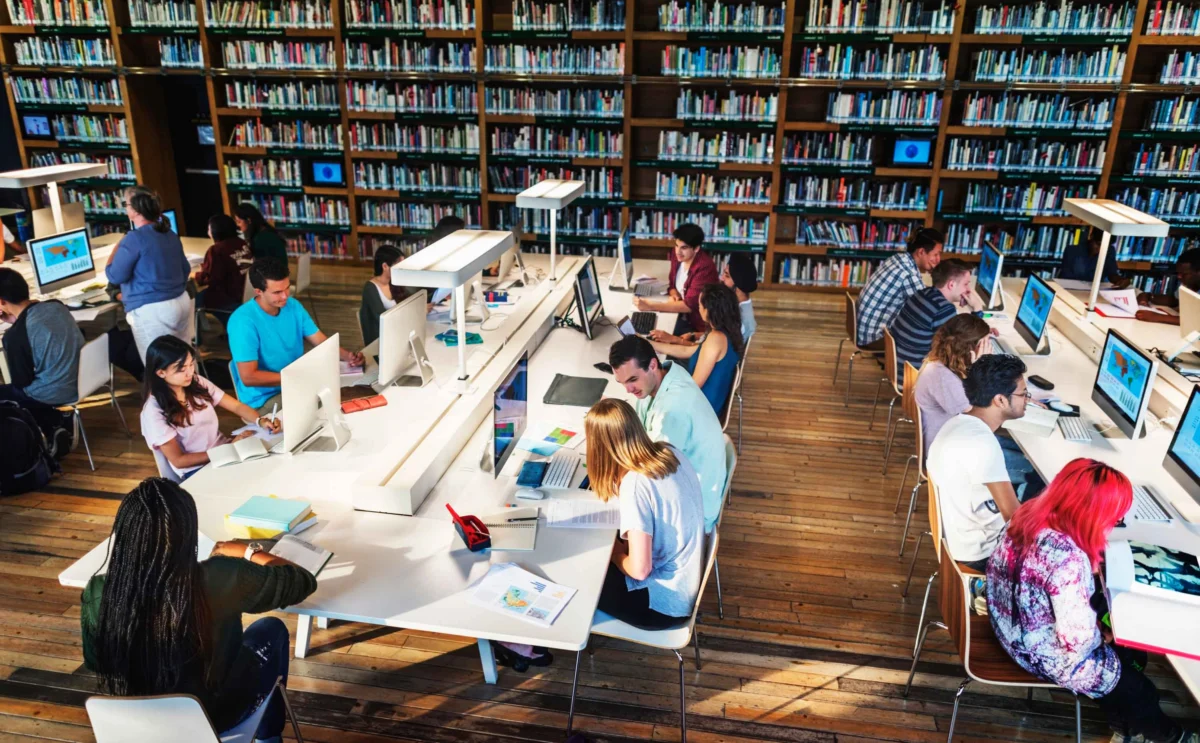 Students work at computers and tables in a large library with wooden floors and bookshelves filled with books.