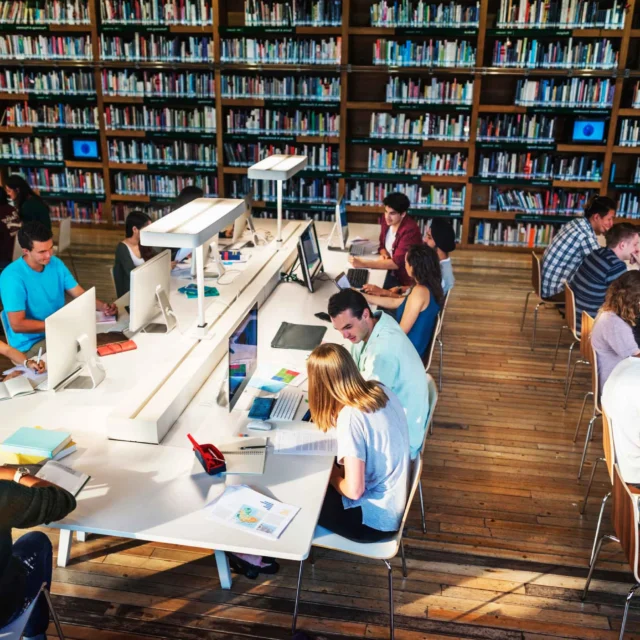 Students work at computers and tables in a large library with wooden floors and bookshelves filled with books.