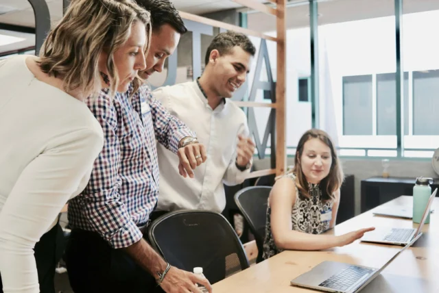 Four people gather around a desk with laptops, actively discussing and looking at a computer screen in a modern office setting.