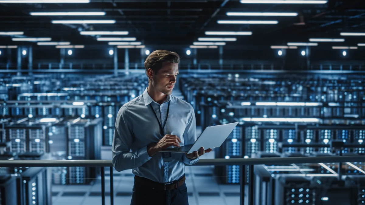 A man stands in a large, modern data center, using a laptop and surrounded by rows of illuminated server racks.
