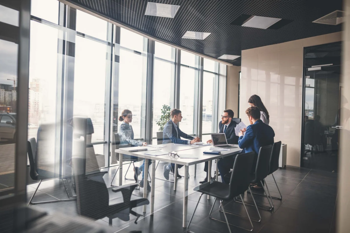 Five people in business attire sit and stand around a conference table in a modern office with large windows and city views, engaged in a meeting or discussion.