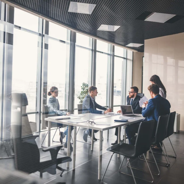 Five people in business attire sit and stand around a conference table in a modern office with large windows and city views, engaged in a meeting or discussion.