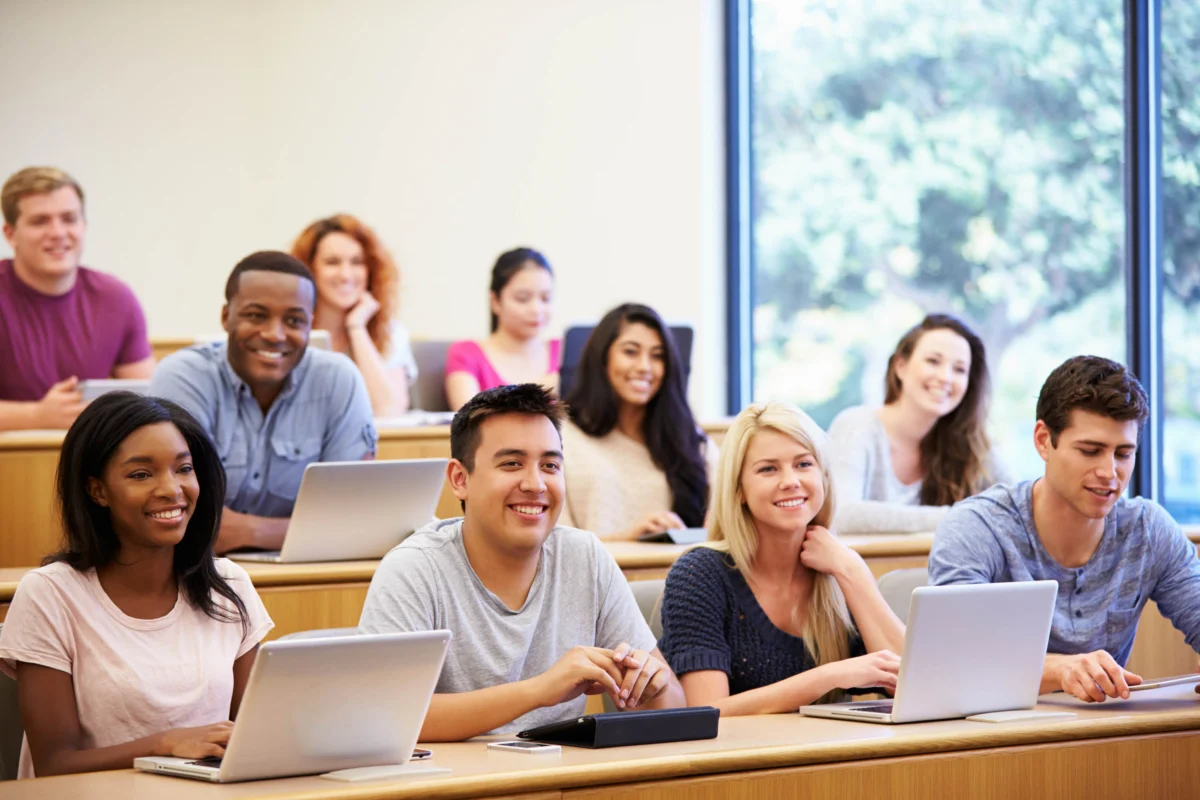 Students sit at desks with laptops and notebooks in a bright classroom, smiling and facing forward, with large windows in the background.