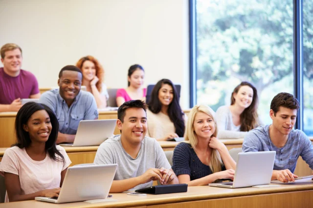 Students sit at desks with laptops and notebooks in a bright classroom, smiling and facing forward, with large windows in the background.