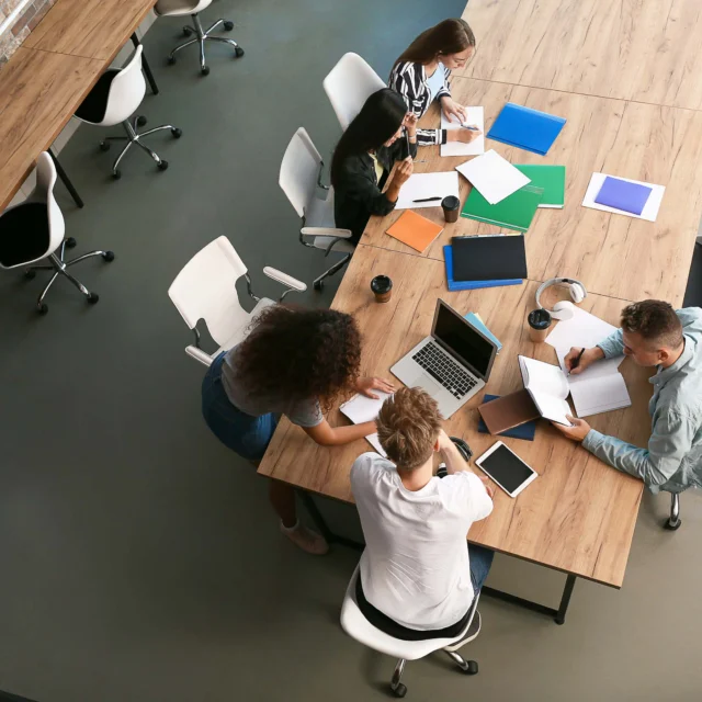 Four people sit around a conference table with laptops, notebooks, and folders, engaged in discussion and working in a modern office setting.
