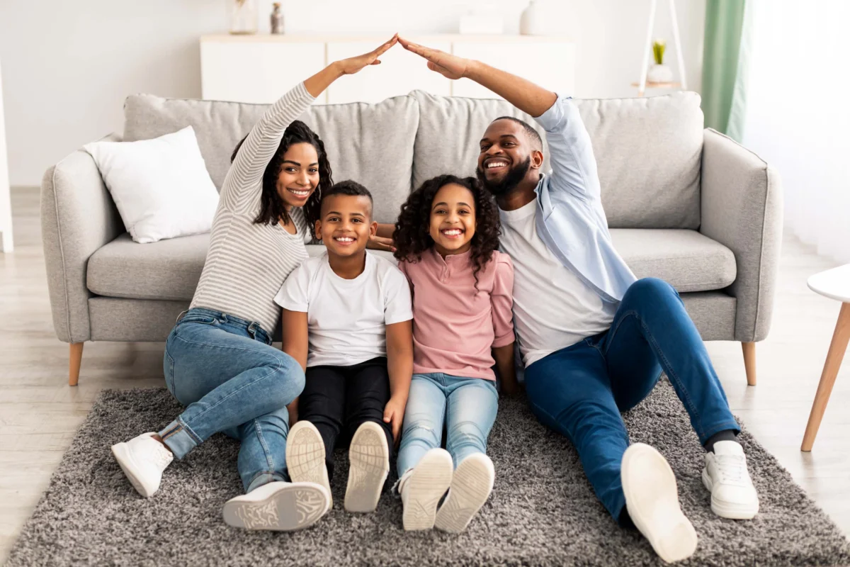 Four people, two adults and two children, sit on a rug in front of a sofa. The adults form a roof shape with their arms over the children. All are smiling.