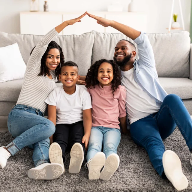 Four people, two adults and two children, sit on a rug in front of a sofa. The adults form a roof shape with their arms over the children. All are smiling.