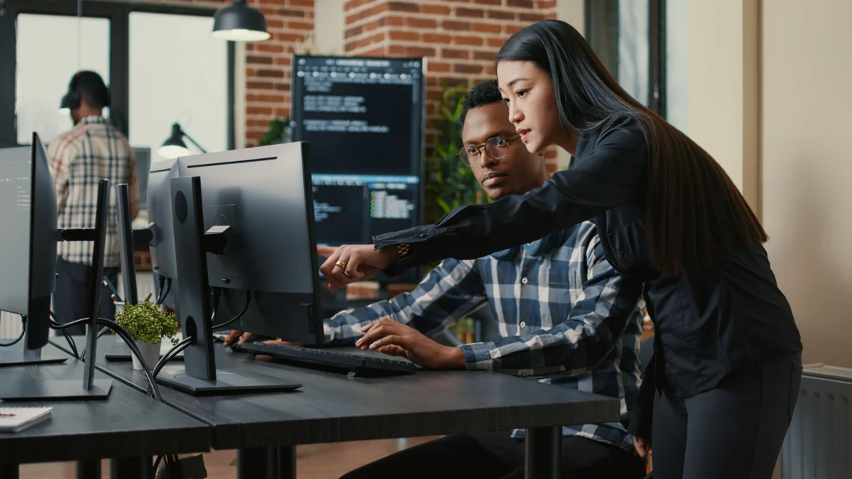 Two people work at a desk with computers; one is seated typing while the other stands and points at the monitor. Code is visible on nearby screens in a modern office setting.
