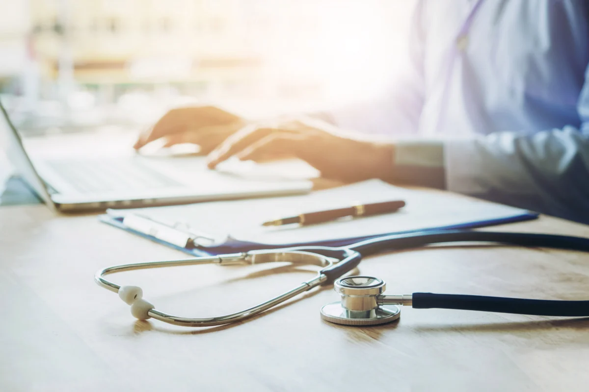 A stethoscope, clipboard, and pen rest on a desk while a person in a dress shirt works on a laptop in the background.