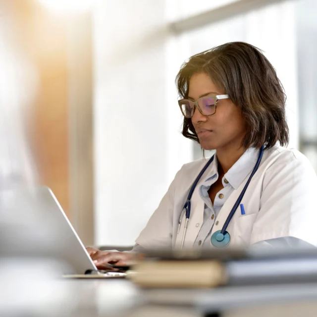 A doctor wearing a white coat and stethoscope sits at a desk, typing on a laptop in a bright office.