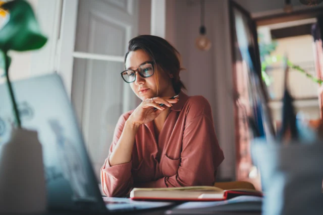 Woman wearing glasses sits at a desk looking at a laptop screen with an open notebook nearby in a softly lit room.
