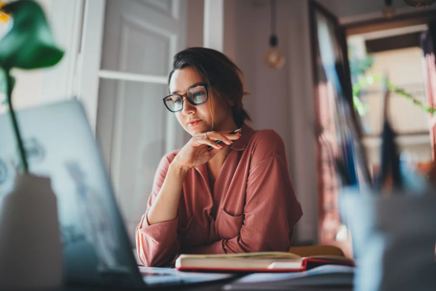 Woman wearing glasses sits at a desk looking at a laptop screen with an open notebook nearby in a softly lit room.