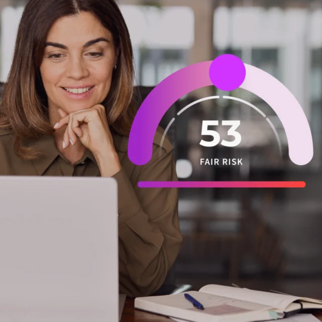 A woman sits at a desk using a laptop. Next to her, a graphic displays a risk score of 53 labeled “Fair Risk.” Open notebooks and a pen are visible on the desk.