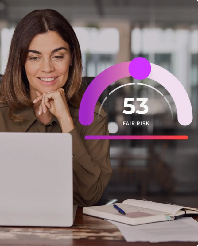 A woman sits at a desk using a laptop. Next to her, a graphic displays a risk score of 53 labeled “Fair Risk.” Open notebooks and a pen are visible on the desk.
