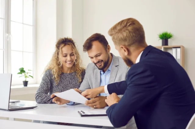 Three people sit at a desk in an office, reviewing documents together and smiling. A laptop and clipboard are on the table.