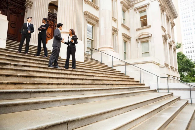 Four people in business attire stand on the steps outside a large, classical-style building, engaged in conversation.
