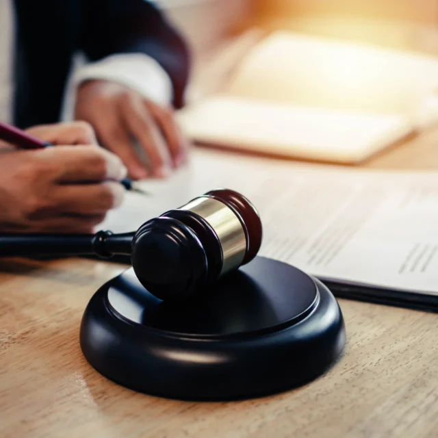 A person in a suit signs documents at a desk with a wooden gavel and legal papers in the foreground.