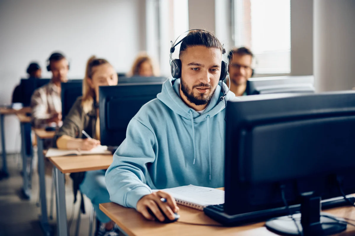 A group of students sits at desks using desktop computers; the student in the foreground wears headphones and a blue hoodie, focused on his screen.