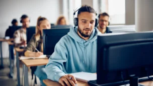 A group of students sits at desks using desktop computers; the student in the foreground wears headphones and a blue hoodie, focused on his screen.