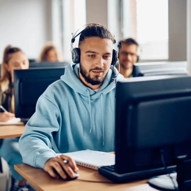 A group of students sits at desks using desktop computers; the student in the foreground wears headphones and a blue hoodie, focused on his screen.