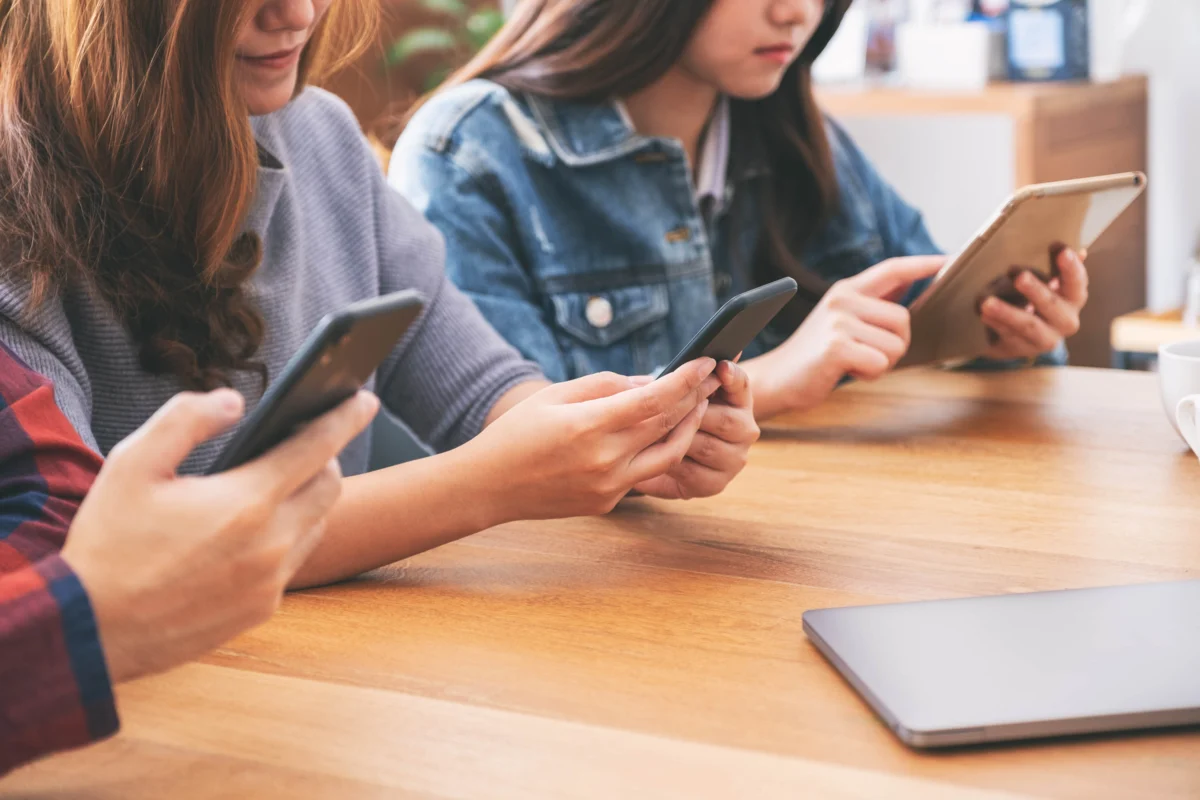 Three people sit at a wooden table using smartphones and a tablet, with a closed laptop nearby.