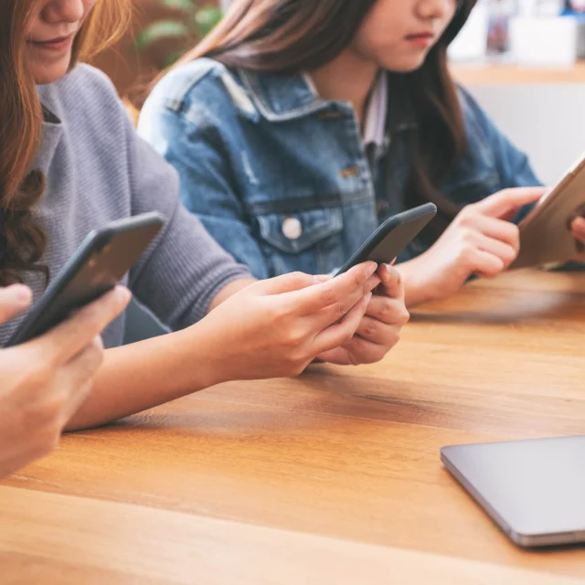 Three people sit at a wooden table using smartphones and a tablet, with a closed laptop nearby.