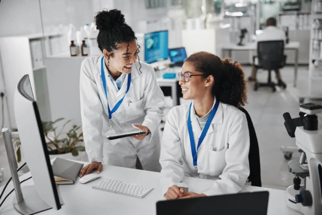 Two scientists in lab coats and ID badges smile and talk at a computer workstation in a modern laboratory setting.