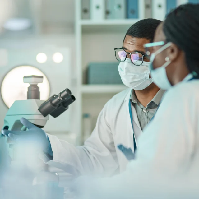 Two scientists wearing lab coats and face masks look at data on a computer screen next to a microscope in a laboratory setting.