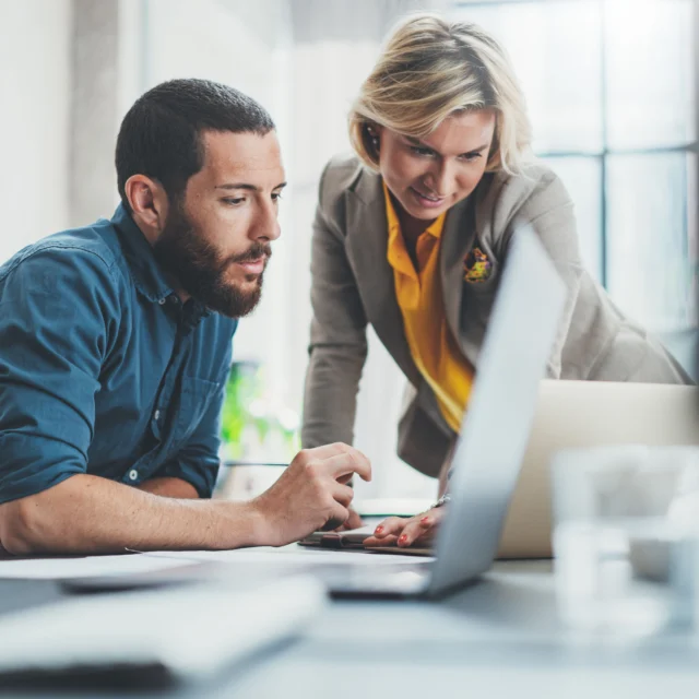 Two people in business attire look at a laptop screen together in an office setting, appearing to discuss or review work.