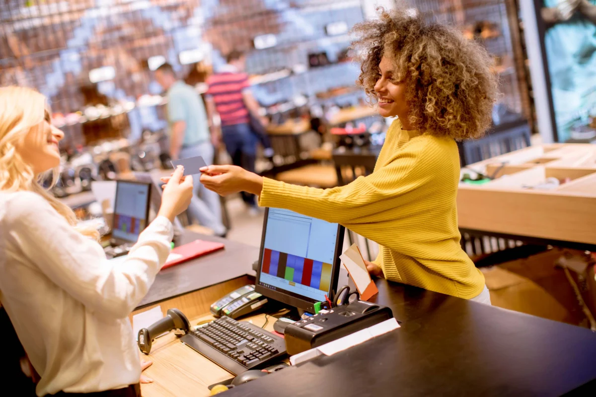 A cashier in a yellow sweater hands a payment card back to a customer at a retail store counter, with computers and shopping items visible.