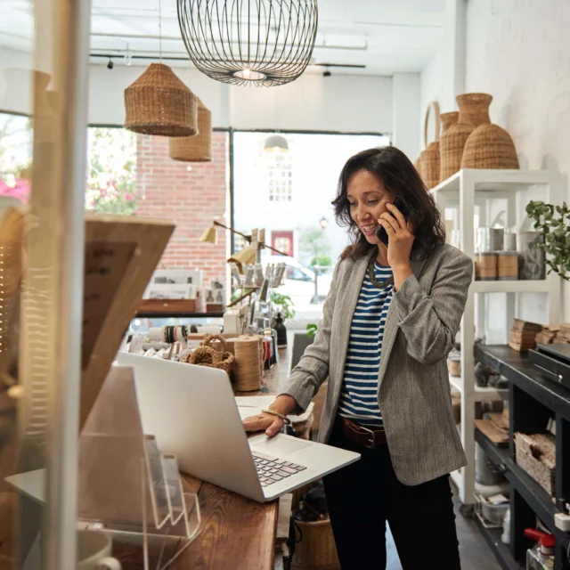 A woman stands in a boutique shop, using a laptop and talking on her phone, surrounded by woven baskets and decorative items.
