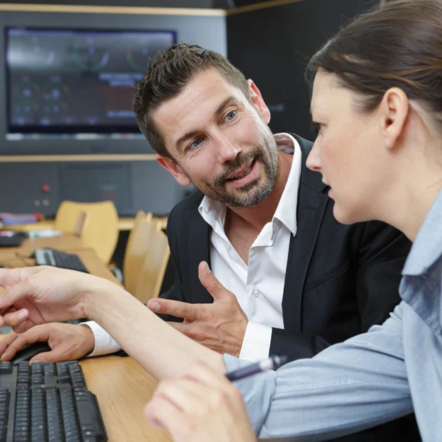 A man and a woman sit at a desk in front of a computer, engaged in a discussion while looking at the monitor.