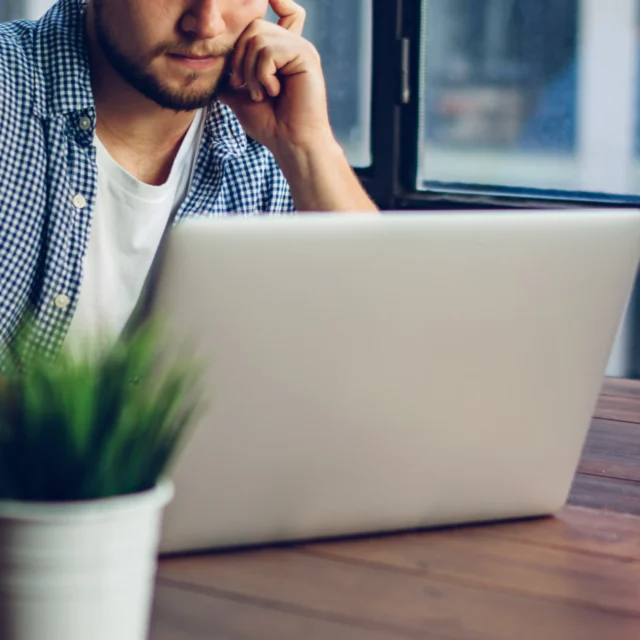 A person sits at a wooden desk using a laptop, with a potted plant and a lit lightbulb lamp nearby, next to a large window.